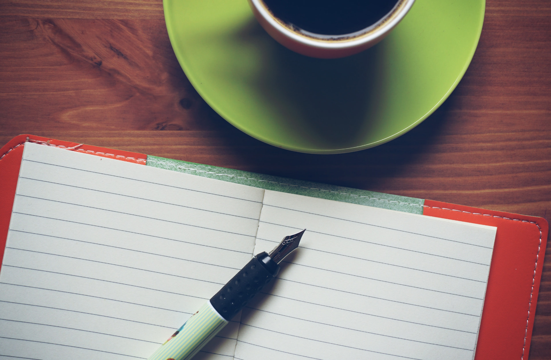 Un carnet ouvert et un stylo plume sur une table en bois, près d’une tasse verte